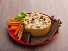 a bowl of dip surrounded by crackers, carrots and celery on a red plate