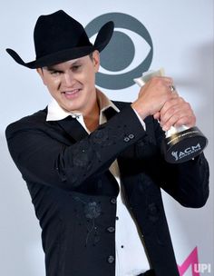 a male in a black jacket and hat holding an award for best country album at the american music awards