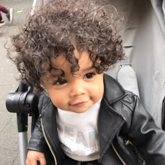 a small child with curly hair sitting in a stroller wearing a jacket and white t - shirt