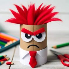 a toilet paper roll with a red hair and tie on it, sitting next to colored pencils