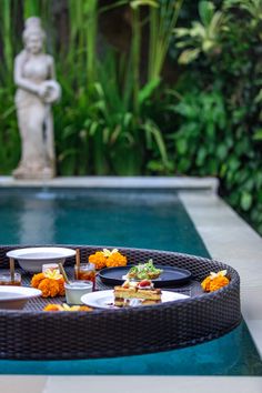 an outdoor table with food on it next to a swimming pool in the middle of a garden