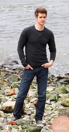 a young man standing on rocks by the water with his hands in his pockets and looking at the camera