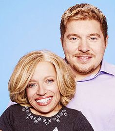 a man standing next to a woman in front of a blue background wearing a purple shirt