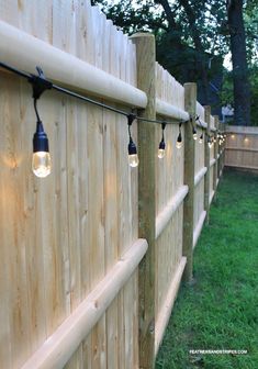 some lights are hanging on the side of a wooden fence with grass and trees in the background