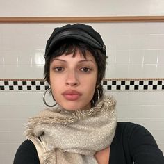 a woman with short hair wearing a hat and scarf in front of a tiled wall