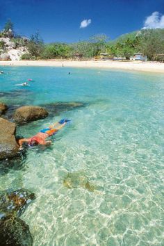 a man swimming in the clear blue water near some rocks and beach people are on the shore