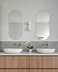 two white sinks sitting on top of a wooden cabinet next to mirrors and a vase with flowers