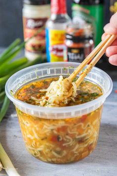 a person holding chopsticks over a bowl of soup with noodles and vegetables in the background