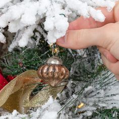 a hand is holding a christmas ornament in front of a snow covered tree