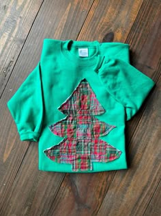 a green shirt with a plaid christmas tree on the front and bottom, sitting on a wooden floor