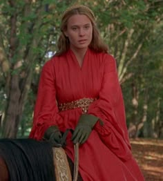 a woman in red riding on the back of a brown and black horse through a forest