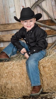 a young boy sitting on top of a hay bale wearing a black cowboy hat