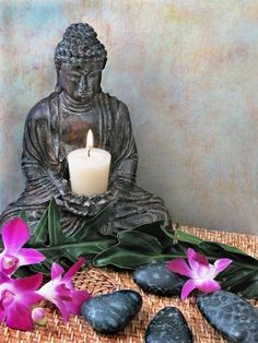 a buddha statue sitting on top of a table next to some flowers and rocks with a lit candle in front of it