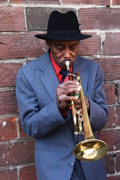 a man in a suit and hat playing a trumpet against a brick wall with his hands