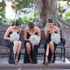 three women sitting on a bench looking at their cell phones while holding bouquets of flowers