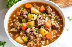 a white bowl filled with stew and vegetables on top of a table next to bread