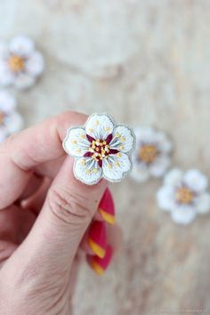 a hand holding a flower shaped brooch in front of white and yellow flowers on the ground