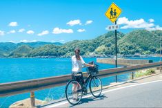 a woman is riding her bike on the road by the water with mountains in the background