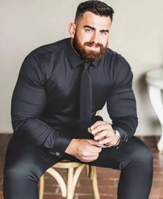 a man with a beard sitting on a chair wearing a black shirt and tie, looking at the camera