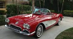 an old red convertible car parked in front of a house with trees and bushes behind it