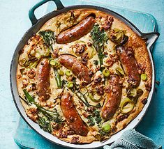 a pizza with sausages and vegetables in a pan on a blue tablecloth next to a pair of oven mitts