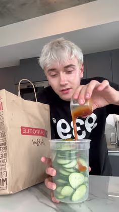 a man is pouring something into a jar with cucumbers in it and a brown bag behind him