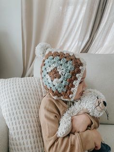 a little boy sitting on a couch holding a teddy bear wearing a crocheted hat