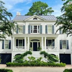 a large white house with black shutters on the front and second story, surrounded by greenery