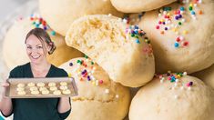 a woman holding a tray with cookies and sprinkles next to a pile of doughnuts