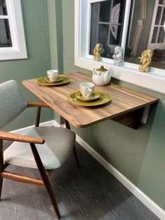 a wooden table with two plates and cups on it next to a chair in front of a window