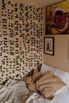 an unmade bed with white sheets and green leaves on the wall behind it is surrounded by string lights