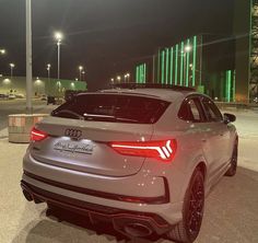 the rear end of a silver car parked in a parking lot at night with lights on