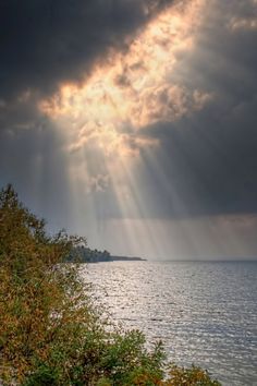 the sun shines through dark clouds over the water and trees in front of it