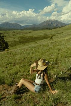 a woman sitting on the ground wearing a cowboy hat and jeans shorts with mountains in the background