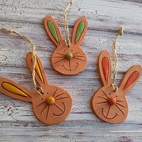 three clay bunny ornaments hanging from twine on wooden planks, one with ears painted orange and the other green