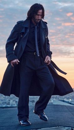 a man with long hair wearing a trench coat and black shoes standing on top of a roof