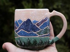 a hand holding a coffee mug with mountains painted on the side and trees in the background