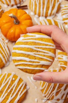 a hand holding a pumpkin cookie with white icing on it and several mini pumpkins in the background