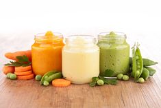 three jars filled with different types of food on top of a wooden table next to carrots and peas