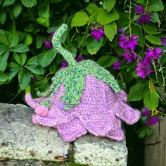 a knitted turtle sitting on top of a stone wall next to purple and green flowers