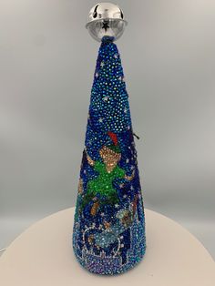 a blue glass christmas tree with a silver bell on it's top and the base is made out of beads