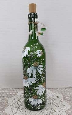 a green glass bottle with white flowers painted on the front and sides, sitting on a doily