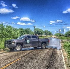 a black truck driving down a wet road next to green grass and trees with blue sky in the background