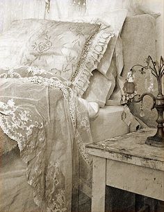 a bed with white sheets and lace on it's headboard, next to a small side table