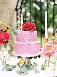 a pink cake sitting on top of a table next to two candles and some flowers