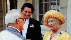 an older woman in a yellow dress and hat talking to two younger men wearing suits