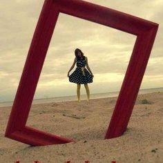 a woman standing on top of a sandy beach next to an empty red frame in the sand