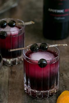 two glasses filled with red liquid and black olives next to an orange on a wooden table