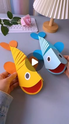 two paper fish are being held by someone's hand on a table next to a lamp