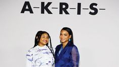 two women standing next to each other in front of a wall with the words akri - is on it